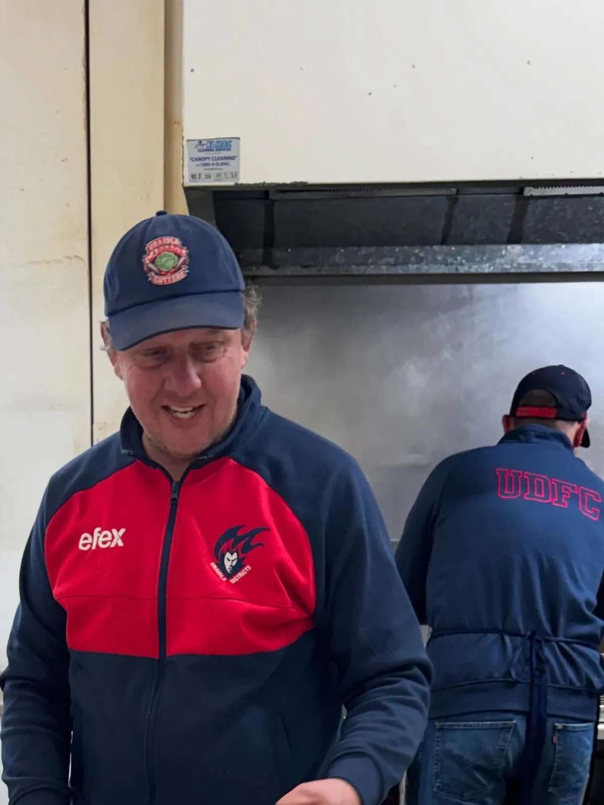 A volunteer in a club jacket and cap cooking at the clubrooms barbecue